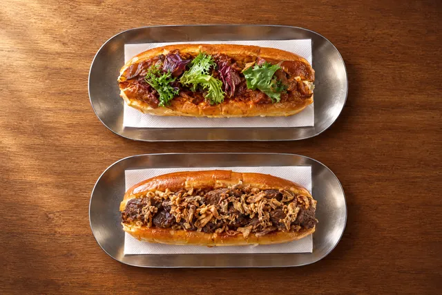 Dos panes brioche rellenos en bandejas de acero sobre madera: uno con carne desmigada en salsa y brotes verdes, otro con carne y cebolla crujiente.
