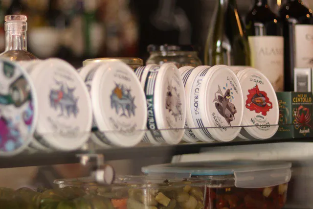 Latas de conservas La Curiosa en la vitrina, con botellas de vino detrás.