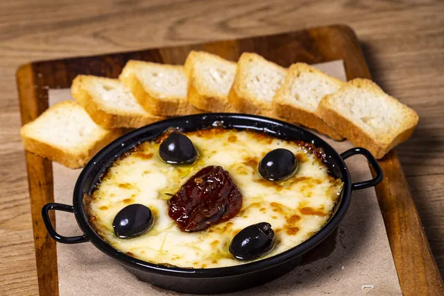 Provoleta fundida en su sartén, con aceitunas negras y tomate seco, y pan tostado alrededor.