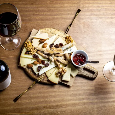 Tabla de quesos con frutos secos, picos y mermelada, entre copas de vino tinto.