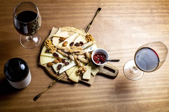 Tabla de quesos con frutos secos, picos y mermelada, entre copas de vino tinto.